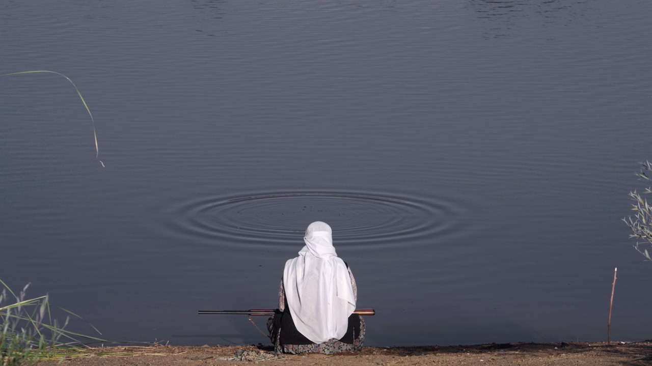 A woman sitting at a lake, rifle in her lap